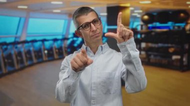 Gözlüklü adam ezik jestler yapıyor ve kameraya bakıyor. Parlak ışıklandırılmış spor salonu, spor aletleri ve modern tasarım..