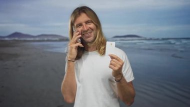 Sarışın adam akıllı telefonu kulağına tutuyor, gülümsüyor ve güneşli kumlu sahilde kredi kartı gösteriyor; seyahat planlama heyecanı.