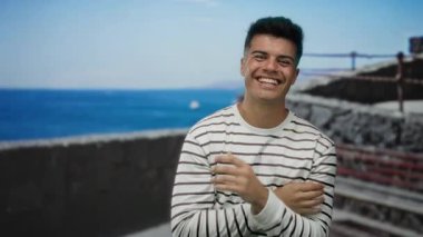 Genç adam, güneşli bir günde okyanus geçmişiyle deniz kenarında gülümsüyor.