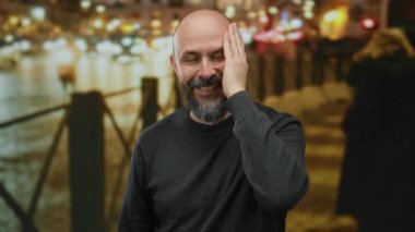 Sakallı bir adam gece şehir sokağında gülüyor. Arka planda bulanık ışıklar var. Canlı ve neşeli bir şehir manzarası yaratıyor..