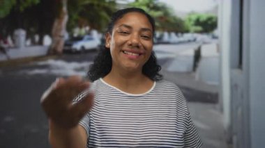 Çizgili tişörtlü gülümseyen kadın, güneşli bir şehir caddesinde kameraya doğru elini uzatıyor..