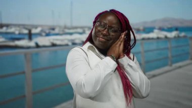 Rıhtımda gözleri kapalı bir kadın uyku jesti taklidi yapıyor, gözlük ve örgü takıyor, arka planda kayıklar ve deniz var..