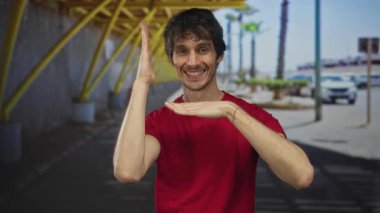 Kırmızı gömlekli, gülümseyen ve çerçeveli görünümlü bir adam, güneşli şehir caddesinde sarı kirişlerin altında elleri ile; neşe.