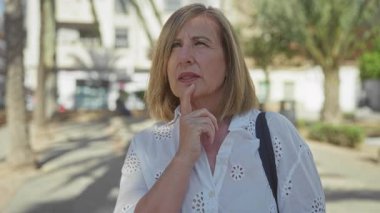 Beyaz bluzlu kadın, güneşli şehir caddesinde palmiye ağaçlarıyla kaplı yukarı bakarken çenesine dokunuyor..