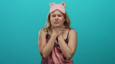 Mavi arka planda pembe gözlü, gecelikli genç sarışın kadın boğazını tutuyor ve rahatsızlık gösteriyor..