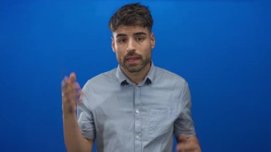 Genç İspanyol adam elini sallıyor ve açık mavi gömlekli, açık mavi arka planda ağzını gösteriyor..