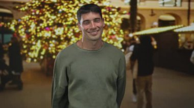 Sokakta aydınlanmış yılbaşı ağacının önünde elleri arkadan sıkıca bağlanmış gülümseyen İspanyol genç adam; sevinç sıcaklığı.