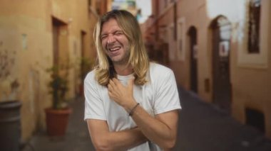 Güneşli caddede çıplak kollarıyla tişörtünü tutan bir adam. Yaşlı binalar ve saksı bitkileriyle çevrili. Acı..