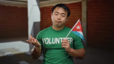 Yeşil gönüllü gömlekli genç adam kapalı inşaat sahasında Küba bayrağı taşıyor, kültürel çeşitlilik ve toplum ruhunu endüstriyel bir ortamda taşıyor..