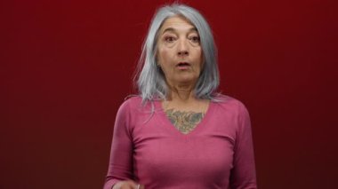 Gri saçlı bir kadın, canlı kırmızı bir arkaplan karşısında ağzını kapatır. Pembe gömlek giyer, duygularını aktarır ve izolasyon yapar..