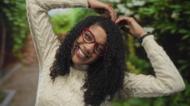 Orman yolunda elleri yukarıda kıvırcık saçlı ve kırmızı gözlüklü, kablo örülü süveter giyen bir kadın kalp şeklini alıyor..