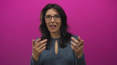 Orta yaşlı İspanyol kadın, parlak mavi bir bluz giyiyor. Gözlük takıyor, elleri açık, ağzı açık, pembe stüdyoda gözleri açık..