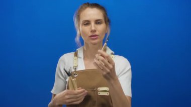 Önlük takan kadın ressam mavi arka planda boya fırçası tutuyor, odaklanmış resim sahnesini izole bir manzarayla tasvir ediyor.