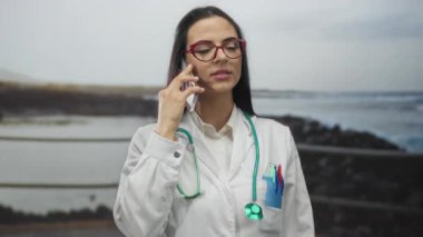 Üniformalı kadın doktor, deniz kenarında steteskopla konuşuyor. Açık havada, bulutlu havada, sakin, profesyonel bir tavır sergiliyor..