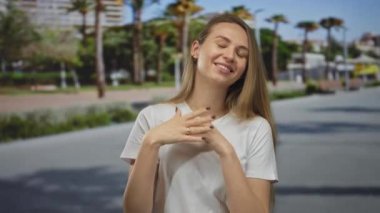 Güneşli bir şehir caddesinde arka planda palmiye ağaçlarıyla gülümseyen bir kadın, gündüz vakti mutluluğu ve şehir hayatını yakalıyor..