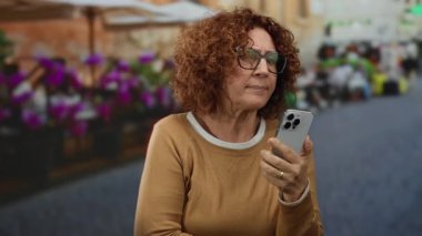 Açık hava restoranındaki terasta akıllı telefonlu bir kadın hareketli şehir ortamında canlı çiçeklerle huzurlu bir anı yakalıyor..