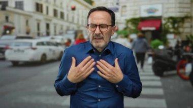 Şehrin işlek caddesinde yürürken mavi tişörtlü adam üşümüş görünüyor. Etrafı bulanık yaya ve araçlarla çevrili.