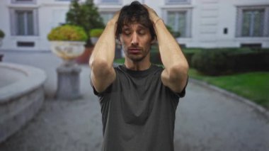 Koyu gri gömlekli genç adam, bir şehir caddesinde beyaz bir binanın önünde ellerini başına bastırır; endişe.