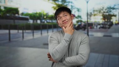 Gri kazaklı genç adam, bulanık şehir arka planı ve yemyeşil ile çevrili hareketli bir şehir caddesinde düşünceli bir şekilde poz veriyor..