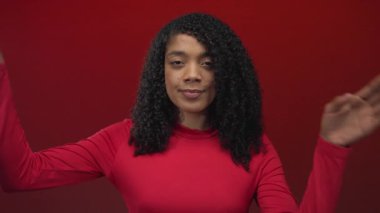 Kırmızı stüdyodaki Afrikalı Amerikalı kadın ellerini tutarak neşeli bir hareketle yüzünü çerçeveliyor..