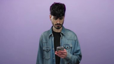 Akıllı telefonlu genç adam, izole edilmiş mor arka plana karşı hayal kırıklığına uğramış görünüyor. Modern dijital hayal kırıklığını, şık bir görünüme ve derin derin düşüncelere sahip bir duruşla ifade ediyor..