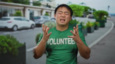 Yeşil gönüllü tişörtü giyen genç bir adam işlek bir şehirde, şehir merkezinde arabaları ve yeşillikleri olan duygusal hareketler yapıyor..