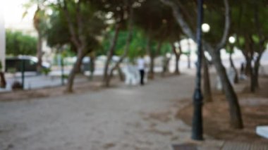 Arka planda bulanık ağaçlar, patika, çözülmüş insan ve uzaktaki beyaz park banklarının olduğu açık hava sahnesi