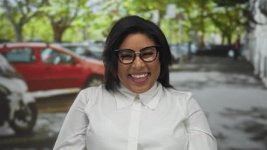 Arka planda arabaları olan, gözlüklü ve beyaz gömlekli, mutluluğu ve şehir yaşam tarzını temsil eden şehir sokaklarında gülümseyen bir kadın..