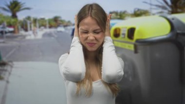 Beyaz bluzlu sarışın kadın şehir caddesinde elleri kulaklarıyla kulaklarını kapatır; sıkıntı stresi bunaltır.