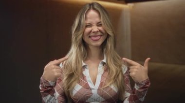 Modern bir stüdyoda dururken kafasını gösteren uzun sarı saçlı ve gülümseyen yüzlü bir kadın. Kendine güvenen bir oyuncu..