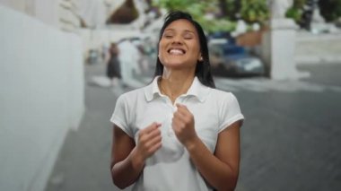 Siyah saçlı bir kadın güneşli bir sokakta beyaz gömlek giyerek neşeyle gülümsüyor, şehir ortamında mutluluk ve enerjiyi yakalıyor..