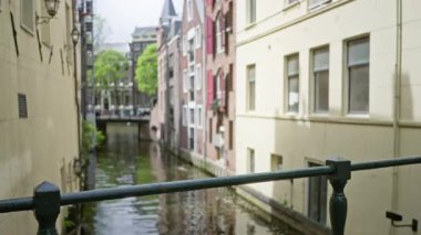 Amsterdam 'daki bulanık kanal manzarası canlı yeşillikle büyüleyici binaları ve sakin su yolunu gösteriyor. Şehir keşfi ve huzur dolu şehir manzarasını taşımak için mükemmel..