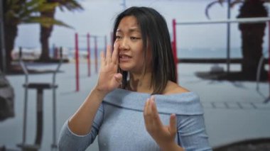 Arka planda palmiye ağaçları olan açık hava spor salonunda bağıran bir kadın mavi bir kazak giyiyor ve heyecan veya motivasyon belirtileri gösteriyor..