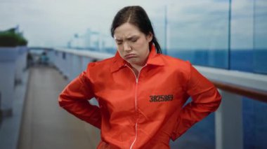 Turuncu hapishane tulumu giyen bir kadın, açık bir gökyüzünün altında, bir yolcu gemisinin güvertesinde sıkıntılı görünüyor..