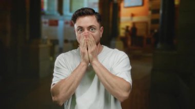 Sakallı genç bir adam şehirde bir caddede duruyor, şaşırdığını ifade ediyor, açık hava ortamında günlük beyaz bir gömlek giyiyor, elleri ağzını kapatıyor..