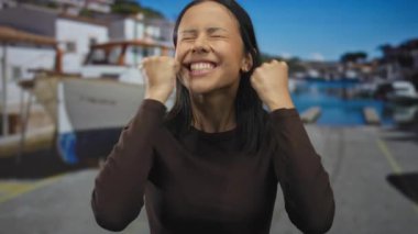 İspanyol kadın deniz kenarında neşeyle kutluyor, manzaralı bir liman ve arka planda tekneler, açık havada mutluluğun ve coşkulu heyecanın özünü yakalıyor..