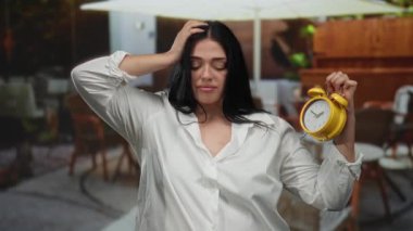 Açık hava restoranının terasında elinde sarı alarmlı saat tutan siyah saçlı genç bir kadın. Sıradan bir ortamda beyaz gömlek giyiyor, yorgun hissediyor..