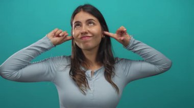 Açık mavi uzun kollu bluz giyen genç İspanyol kadın stüdyoda parmaklarıyla kulaklarını kapatıyor..