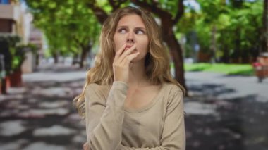 Kadın endişeli bir bakışla eline bastırır ve bir akıllı telefonu kavrar; sarışın genç ziyaretçi aramadan önce endişeli bir şekilde parkın açık alanında durur.