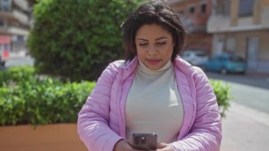 Pembe ceketli genç İspanyol kadın parkta akıllı telefon kullanıyor, doğayı eğlence ve bağlantı için kentsel ortamla harmanlıyor..