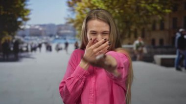 Kadın hareketli bir şehir caddesinde gülüyor ve kamerayı işaret ediyor. Canlı duygular ve şehir enerjisi sergiliyor. Arka planda yoldan geçenler ve yol boyunca ağaçlar var..