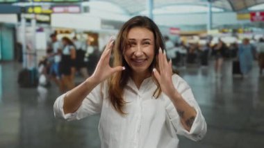 Kapalı bir havaalanı terminalinde gülümseyen genç bir kadın, arka planda canlı bir atmosfer ve çeşitli gezgin aktiviteleri sergiliyor..