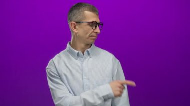 Gözlüklü beyaz bir adam, parlak mor bir arka plana karşı, çizgili bir gömlek giymiş, merak ya da endişeyle bakıyor..