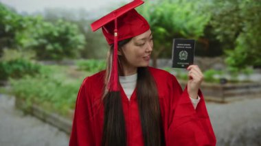 Kırmızı mezuniyet elbisesi giymiş bir kadın parkta İtalyan pasaportu taşıyor. Çinli mezunlar için küresel seyahat olanaklarını ve başarıyı simgeliyor..