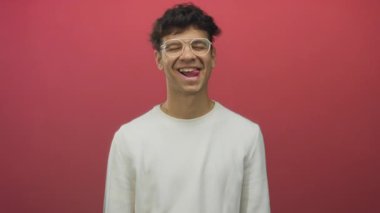 Yuvarlak gözlüklü ve beyaz gömlekli adam kırmızı stüdyoda dilini çıkarıp gözlerini kapatıyor..