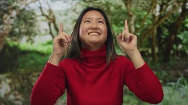 Açık bir gökyüzünün altında ağaçlarla kaplı yeşil bir parkı işaret eden kırmızı kazaklı bir kadın açık hava ortamında iyimserlik ve neşe öneriyor..