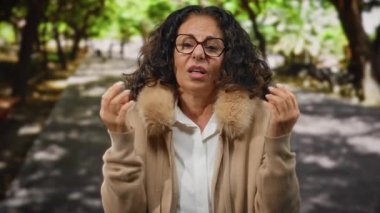 Kıvırcık saçlı ve gözlüklü olgun bir kadın, verimli yeşilliklerle çevrili güneşli bir parkta huzurlu bir anın tadını çıkarıyor..