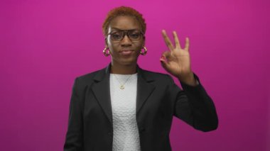 Gözlük ve halka küpe takan bir kadın, parlak pembe bir stüdyo geçmişiyle el işareti yapıyor. Onaylandı..