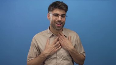 Gözlüklü, bej gömlekli, göğüs debriyajlı, parlak mavi bir stüdyoda genç bir İspanyol..