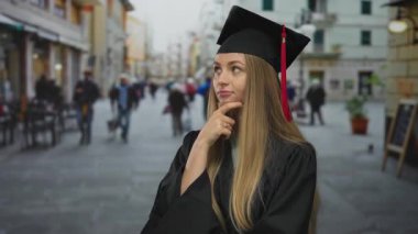 Mezuniyet kepi ve cübbesi takmış genç bir kadın yoğun bir şehir caddesinde düşünceli bir şekilde duruyor, akademik başarıyı açık hava ortamında kentsel yaşamla harmanlıyor..